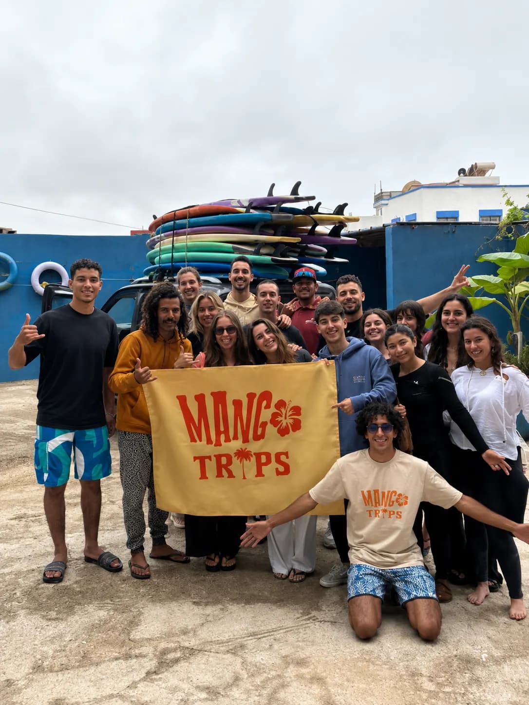 El grupo posa con la bandera de Mangotrips en el surfcamp, con las tablas en el rack detrás